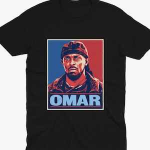 May include: A black short-sleeved t-shirt featuring a graphic print of a man wearing a cap. The design uses a red and blue colour palette with the name OMAR printed in light blue capital letters at the bottom of the rectangular frame.