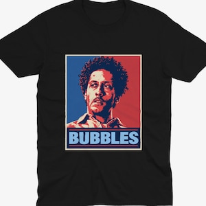 Może przedstawiać: Czarny t-shirt z krótkim rękawem z nadrukiem graficznym przedstawiającym osobę z kręconymi włosami. Projekt wykorzystuje czerwono-niebieską kolorystykę z napisem BUBBLES wydrukowanym jasnoniebieskimi wielkimi literami u dołu prostokątnej ramki.