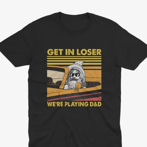 Könnte beinhalten: Ein schwarzes Kurzarm-T-Shirt mit einer Grafik eines bärtigen Zauberers mit Sonnenbrille in einem gelben Cabriolet. Der Text GET IN LOSER WE'RE PLAYING D&D ist vor einem Hintergrund aus horizontalen gelben und roten Streifen zu sehen.