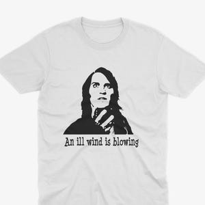 Peut inclure: Un t-shirt blanc à manches courtes avec une impression graphique noire représentant une personne aux cheveux longs tenant une main devant son visage. Sous le graphique, le texte indique "An ill wind is blowing" en police noire.