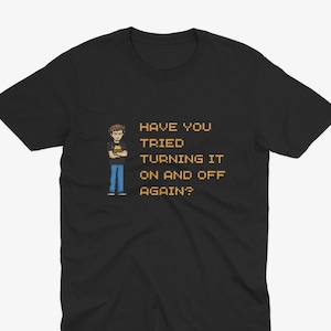May include: Black t-shirt with the text "HAVE YOU TRIED TURNING IT ON AND OFF AGAIN?" in yellow pixelated font. A pixelated image of a person is on the left side of the shirt.