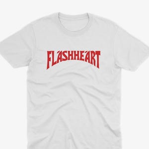 May include: A white short-sleeved t-shirt featuring the word FLASHHEART printed in a bold, red, arched font across the center of the chest. The garment has a crew neckline and a standard fit, showcasing a clean design with high contrast.