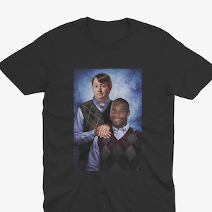 Peut inclure: Un t-shirt noir avec une photo de deux hommes portant des pulls. L'homme à gauche porte une chemise bleue et un pull à motifs marrons et blancs. L'homme à droite porte une chemise bleue et un pull à motifs noirs et blancs. Ils sourient tous les deux et regardent la caméra.