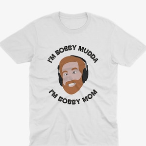 May include: White t-shirt with a cartoon illustration of a man with a beard and headphones. The text "I'm Bobby Mudda I'm Bobby Mom" is printed on the shirt.