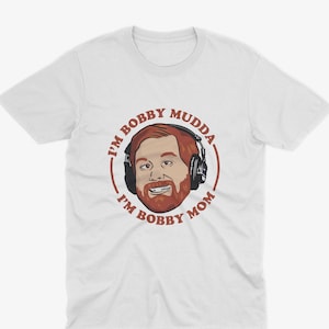 May include: White t-shirt with a red circle graphic featuring a cartoon man with a beard and headphones. The text inside the circle reads "I'm Bobby Mudda I'm Bobby Mom."