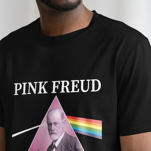 May include: Black t-shirt with a white graphic of a man's face in a pink triangle with a rainbow stripe underneath. The text "PINK FREUD" is printed above the graphic.