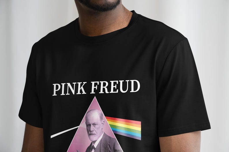 May include: Black t-shirt with a white graphic of a man's face in a pink triangle with a rainbow stripe underneath. The text "PINK FREUD" is printed above the graphic.