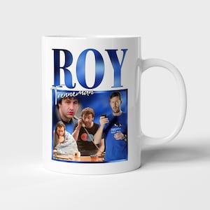 以下が含まれることがあります： 白いセラミックマグカップ。青色の「ROY」の文字と男性の写真がプリントされています。マグカップには取っ手があり、光沢のある仕上げです。画像はコラージュで配置されています。
