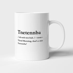 Puede incluir: Una taza de cerámica blanca con texto impreso en negro que dice Tnetennba, seguido de la pronunciación fonética teh-neh-ten-bah, la palabra noun y la frase de ejemplo Good Morning, that’s a nice Tnetennba. La taza tiene un asa.