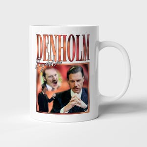 Puede incluir: Taza de cerámica blanca con el nombre "DENHOLM" en letras grandes y estilizadas con efecto degradado. La taza también muestra imágenes de un hombre con traje, con texto adicional "Reynholm". Un asa es visible en el lado derecho.