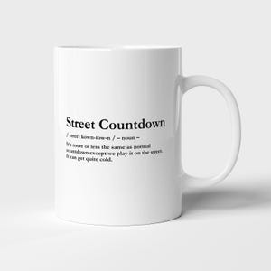 May include: A white ceramic mug featuring black printed text that reads: Street Countdown / street kown-tow-n / - noun - It's more or less the same as normal countdown except we play it on the street. It can get quite cold. The mug has a handle.