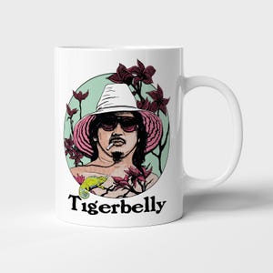 May include: White ceramic mug featuring a graphic illustration of a person wearing sunglasses and a hat, surrounded by flowers and a lizard. The mug has the word "Tigerbelly" printed in black.