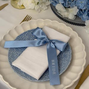 May include: A white fabric napkin folded and tied with a light blue satin ribbon. The ribbon features the word BRIDE printed in white lettering. The napkin rests on a light blue ceramic plate with a floral embossed pattern and scalloped edges.