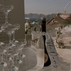 May include: A champagne bottle with a white ribbon bearing the inscription "The Davis's" sits on a clear acrylic surface. A pyramid of champagne glasses is visible, suggesting a wedding. The setting is outdoors, with a scenic backdrop.