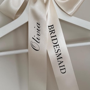 May include: A white clothes hanger featuring a champagne-coloured satin ribbon tied in a bow at the top. The ribbon has the name Olivia printed in a black script font and the word BRIDESMAID printed in a black sans-serif font on the hanging tails.