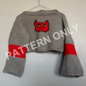 Puede incluir: Un suéter gris recortado con rayas rojas en las mangas y un diseño de gato rojo tejido a crochet en la espalda. El suéter está colgado en una percha blanca.