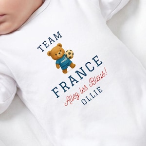 A sleeping baby wearing a white onesie with a teddy bear and text that says "TEAM FRANCE ALEY LES BLUES OLLIE".