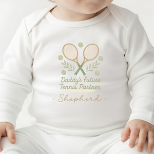 Peut inclure: Body à manches longues blanc pour bébé, orné d'un motif sur le thème du tennis. Le design comprend deux raquettes de tennis croisées, des balles de tennis et le texte "Daddy's Future Tennis Partner - Shepherd" en vert et marron.