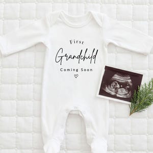May include: A white long-sleeved baby bodysuit featuring the black printed text First Grandchild Coming Soon with a small heart icon. A black and white ultrasound scan is placed next to the bodysuit, accompanied by a small green sprig of foliage.