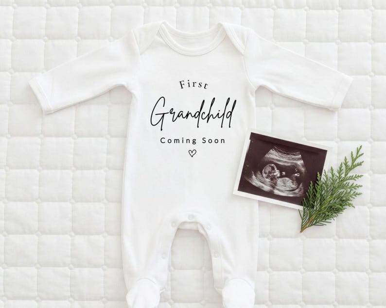 May include: A white long-sleeved baby bodysuit featuring the black printed text First Grandchild Coming Soon with a small heart icon. A black and white ultrasound scan is placed next to the bodysuit, accompanied by a small green sprig of foliage.