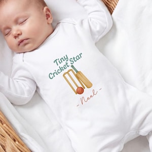 Puede incluir: Un body blanco de bebé con mangas largas con el texto "Tiny Cricket Star" en verde y un gráfico de cricket. El body también incluye el nombre "Neel" en escritura roja. El bebé está acostado en una cesta de mimbre.