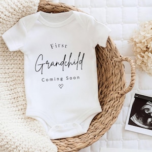 May include: A white short-sleeved baby bodysuit featuring the black printed text First Grandchild Coming Soon with a small heart icon. The garment is made of cotton fabric and includes a snap closure at the bottom for easy diaper changes.