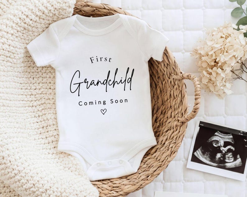 May include: A white short-sleeved baby bodysuit featuring the black printed text First Grandchild Coming Soon with a small heart icon. The garment is made of cotton fabric and includes a snap closure at the bottom for easy diaper changes.