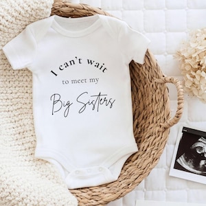 Könnte beinhalten: Ein weißer kurzärmeliger Babybody mit dem schwarzen Aufdruck "I can't wait to meet my Big Sisters" auf der Vorderseite. Das Kleidungsstück hat einen Rundhalsausschnitt und Druckknöpfe im Schrittbereich und liegt in einem geflochtenen Korb.