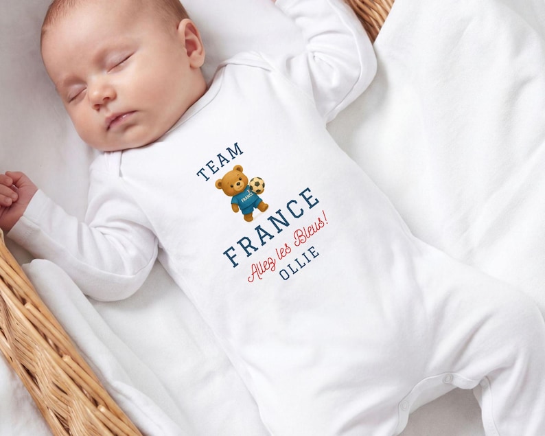 A sleeping baby wearing a white onesie with a teddy bear and text that says "TEAM FRANCE ALEY LES BLUES OLLIE".