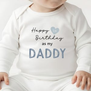 Op de afbeelding: Een witte babybody met lange mouwen en de tekst Happy Birthday 1st as my DADDY in zwarte en blauwe letters. Het ontwerp bevat een klein lichtblauw hartpictogram naast het woord Birthday, met het woord DADDY in een getextureerd blauw lettertype.