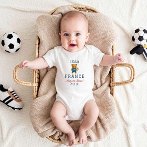 A baby wearing a white onesie with the text "TEAM FRANCE" is lying in a woven basket, surrounded by soccer balls and a stuffed animal.