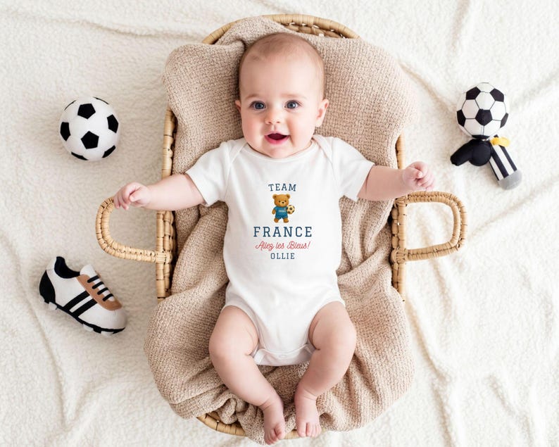 A baby wearing a white onesie with the text "TEAM FRANCE" is lying in a woven basket, surrounded by soccer balls and a stuffed animal.