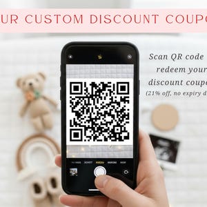 A person is holding a smartphone displaying a QR code, with a teddy bear in the background.
