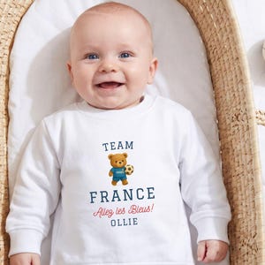 A smiling baby wearing a white sweatshirt with a teddy bear and text printed on it.