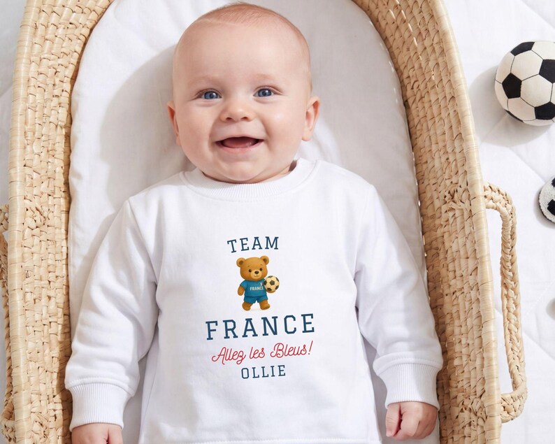 A smiling baby wearing a white sweatshirt with a teddy bear and text printed on it.