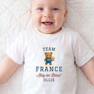 A smiling baby wearing a white t-shirt with a teddy bear and text related to France.