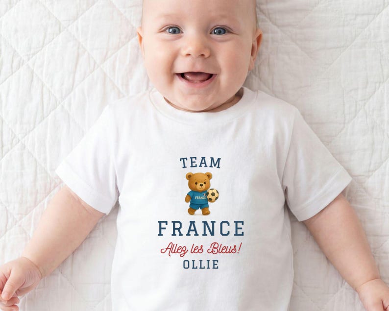 A smiling baby wearing a white t-shirt with a teddy bear and text related to France.