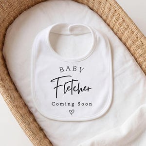 Może przedstawiać: Biały śliniaczek dla dziecka z imieniem "Fletcher" w czarnym skrypcie, wraz ze słowami "Baby" i "Coming Soon" oraz symbolem serca. Śliniaczek umieszczony jest w wiklinowym koszu wyłożonym białym materiałem.
