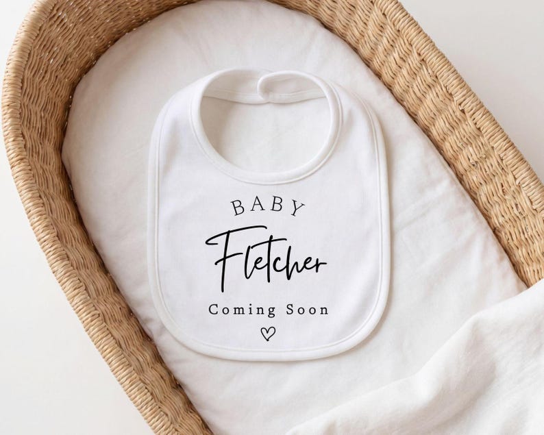Może przedstawiać: Biały śliniaczek dla dziecka z imieniem "Fletcher" w czarnym skrypcie, wraz ze słowami "Baby" i "Coming Soon" oraz symbolem serca. Śliniaczek umieszczony jest w wiklinowym koszu wyłożonym białym materiałem.