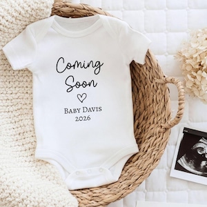May include: A white short-sleeved baby bodysuit featuring the black printed text Coming Soon, a small heart icon, the name BABY DAVIS, and the year 2026. The garment has a round neckline and snap closures at the bottom for easy access.