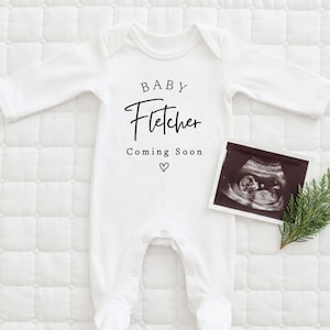 Może przedstawiać: Białe body dla dziecka z napisem "BABY Fletcher Coming Soon" czarnym pismem. Obok body umieszczono zdjęcie USG i gałązkę zieleni na białej, pikowanej powierzchni. Pod tekstem znajduje się symbol serca.