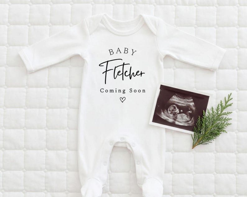 Może przedstawiać: Białe body dla dziecka z napisem "BABY Fletcher Coming Soon" czarnym pismem. Obok body umieszczono zdjęcie USG i gałązkę zieleni na białej, pikowanej powierzchni. Pod tekstem znajduje się symbol serca.