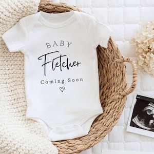 Może przedstawiać: Białe body dla dziecka z imieniem "Fletcher" wydrukowanym czarnym pismem, wraz z "Coming Soon" i symbolem serca. Body umieszczone jest w wiklinowym koszu, w pobliżu znajduje się zdjęcie USG. Biały dzianinowy koc jest w tle.