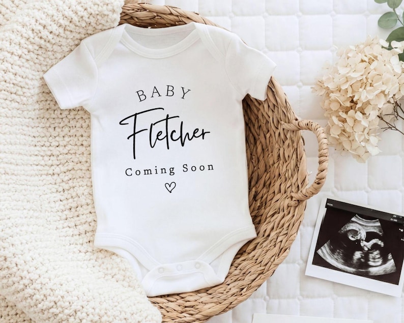 Może przedstawiać: Białe body dla dziecka z imieniem "Fletcher" wydrukowanym czarnym pismem, wraz z "Coming Soon" i symbolem serca. Body umieszczone jest w wiklinowym koszu, w pobliżu znajduje się zdjęcie USG. Biały dzianinowy koc jest w tle.