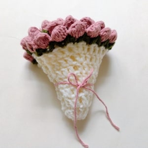 May include: A handmade crochet bouquet featuring pink flowers and green leaves atop a cream-colored, crocheted cone. The bouquet is tied with a pink string. The craftwork is displayed on a white background.
