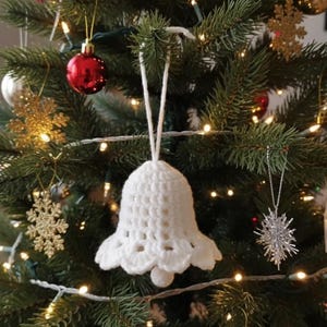 May include: A white, crocheted bell Christmas ornament hanging on a decorated Christmas tree. The tree is adorned with red, gold, and silver ornaments, and string lights. The bell has a scalloped edge and a small ball at the bottom.