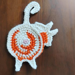 Könnte beinhalten: Ein gehäkelter Katzen-Untersetzer in Weiß und Orange. Die Katze ist zu einer Kugel zusammengerollt, mit dem Schwanz um den Körper gewickelt.