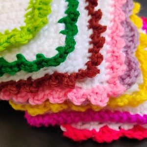 May include: A stack of crocheted squares in various colors, including green, brown, pink, yellow, and red. Each square has a decorative crocheted border.