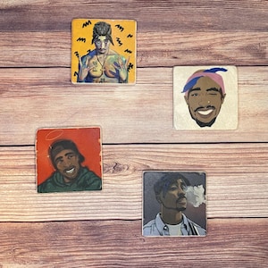 May include: Four square ceramic coasters featuring different images of the rapper Tupac Shakur. The coasters are all white with a black border. The images are all in color and show Tupac in different poses. One coaster shows Tupac with a halo, another shows him with a bandana, another shows him with a cigarette, and the last one shows him with his arms crossed.