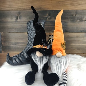 May include: Two Halloween gnomes with long gray beards and tall hats. One gnome wears a black hat with a spider, the other an orange hat with a leaf. A black witch boot is in the background.